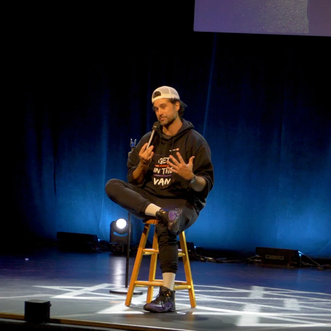 A comedian performing seated on a stool